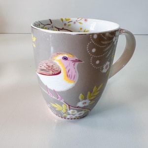 Puede incluir: Taza de cerámica gris con diseño de pájaro y flores. La taza tiene un interior blanco y un asa beige. El pájaro presenta detalles rosas, amarillos y marrones. También hay flores blancas y hojas amarillas.