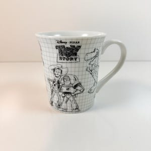May include: White ceramic mug with black and white sketches of Toy Story characters. The mug has a handle and a grid pattern. The Toy Story logo is also visible on the mug's surface.