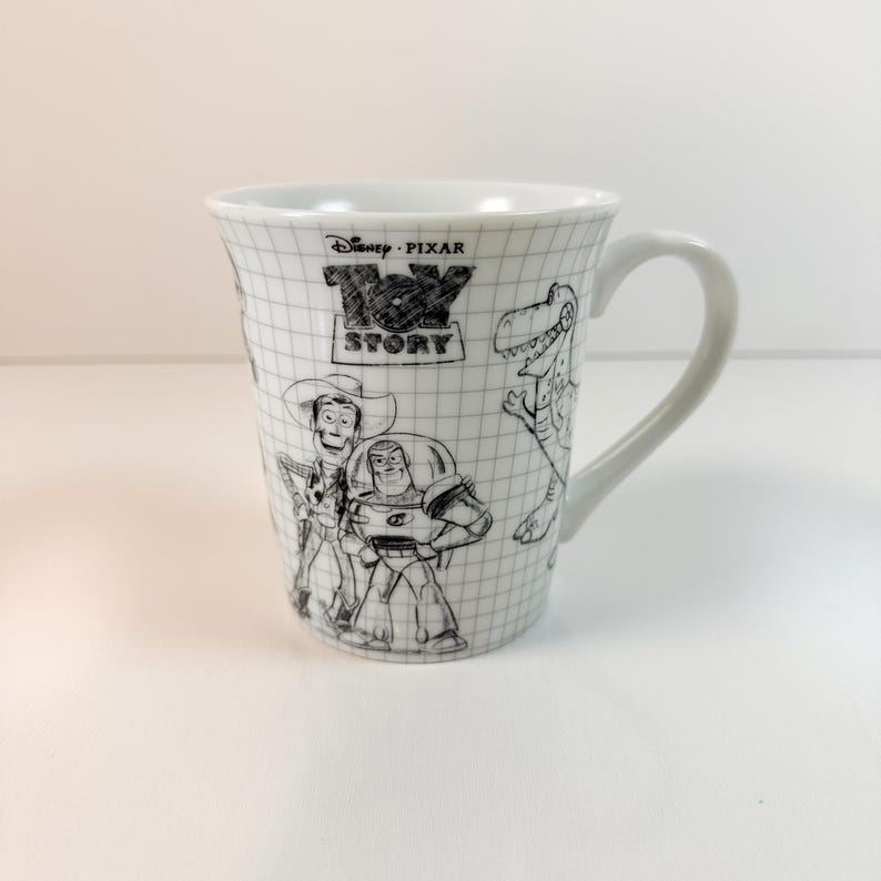 May include: White ceramic mug with black and white sketches of Toy Story characters. The mug has a handle and a grid pattern. The Toy Story logo is also visible on the mug's surface.