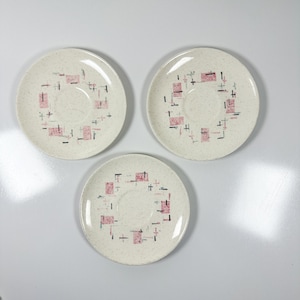 May include: Three off-white saucers with a speckled finish. Each saucer features a central well and a ring of pink and black geometric shapes. The saucers are arranged in a triangular pattern against a white background.