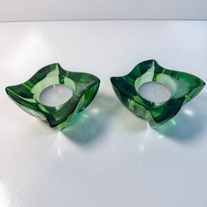 Peut inclure: Deux bougeoirs en verre vert avec un design unique à quatre lobes. Chaque bougeoir contient une bougie chauffe-plat blanche. Le verre a une finition brillante, et l'esthétique générale est moderne et décorative.