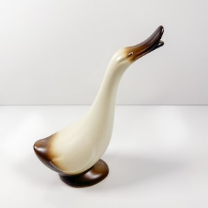 May include: A ceramic goose figurine with a cream-colored body and a brown head and base. The goose is depicted with its head and neck extended upwards, mouth open. The figurine is a decorative item.
