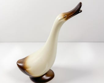 Puede incluir: Figura de ganso de cerámica con cuerpo color crema y cabeza y base marrones. El ganso está representado con la cabeza y el cuello extendidos hacia arriba, con la boca abierta. Es un objeto decorativo.