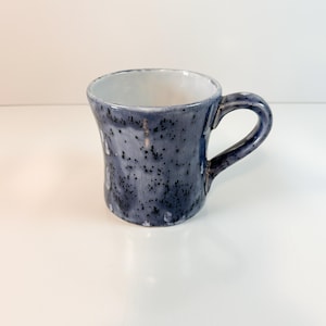 May include: A handmade ceramic mug with a speckled, dark blue glaze. The mug has a rounded body, a curved handle, and a white interior. The mug is approximately 4 inches tall and 3 inches in diameter.