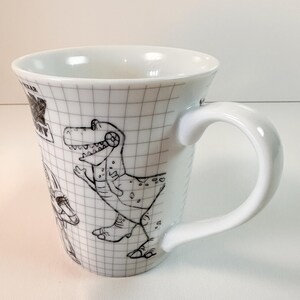 May include: White ceramic mug with a black and white cartoon dinosaur design. The mug has a grid pattern and a curved handle. A collectible item, the mug is suitable for hot beverages.