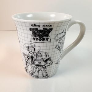 May include: White ceramic mug with black and white illustrations of Toy Story characters. The mug features the Toy Story logo and a grid pattern. The mug has a curved handle.