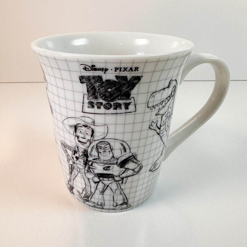 May include: White ceramic mug with black and white illustrations of Toy Story characters. The mug features the Toy Story logo and a grid pattern. The mug has a curved handle.