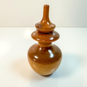 Puede incluir: Un tarro de madera hecho a mano con tapa. El tarro es de color marrón cálido con un acabado brillante. La tapa es una versión más pequeña de la forma del tarro, creando un efecto escalonado. El tarro probablemente se utiliza para almacenamiento o decoración.