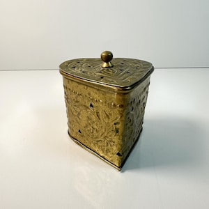 May include: An antique brass heart-shaped box with a lid and a small knob. The box has detailed floral and geometric cut-out patterns. The brass has a warm, aged patina. The box is set against a white background.