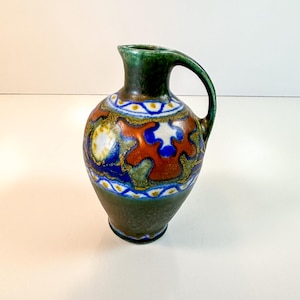 May include: A ceramic pitcher featuring a dark green base and a rounded body. The surface is decorated with a central band of abstract orange, blue, and white shapes, framed by two horizontal borders of white circles with yellow centers.
