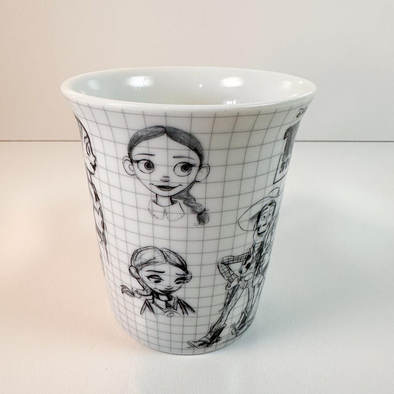 May include: White ceramic mug with pencil sketches of cartoon characters. The mug has a grid pattern. The sketches include a girl with a braid and a cowboy. The mug is cylindrical with a flared top.