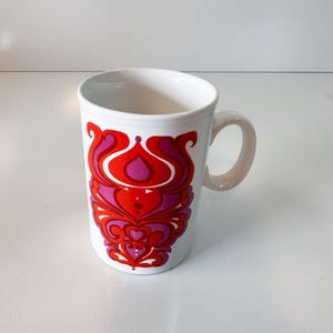 May include: White ceramic mug with a vibrant red and purple floral design. The mug has a curved handle and a cylindrical shape. The design features stylized floral elements in shades of red and purple, creating a bold and eye-catching pattern.