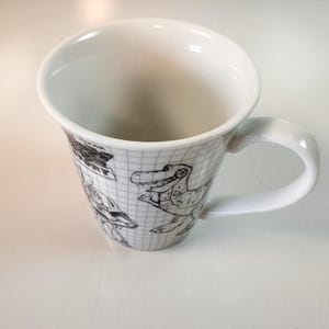 May include: White ceramic mug with dinosaur illustrations. The mug has a grid pattern and a curved handle. The dinosaur drawings are in black ink. The mug is empty and sits on a white surface.