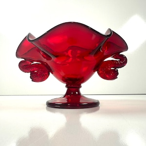 May include: A vibrant red glass bowl with a ruffled rim and decorative, curled accents. The bowl sits on a pedestal base, showcasing a classic design. The glass has a glossy finish, reflecting light.