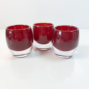 Könnte beinhalten: Drei klare Glas-Teelichthalter mit tiefrotem Oberteil und klarem Boden. Die Teelichthalter sind oval geformt und stehen auf einer weißen Oberfläche.