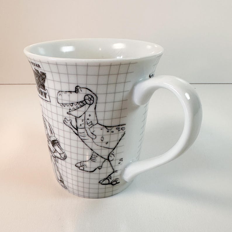 May include: White ceramic mug with black line drawings of cartoon characters on a grid. The mug has a curved handle and a glossy finish. The mug is a Toy Story themed product.