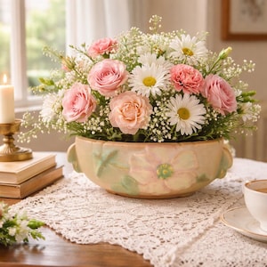 Könnte beinhalten: Ein Blumenarrangement mit rosa Rosen, Gänseblümchen und Nelken in einer dekorativen, hellgelben Keramikschale. Die Schale hat ein Blumendesign und steht auf einer Spitzendecke. Eine brennende Kerze und Bücher sind im Hintergrund zu sehen.