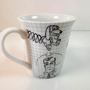 May include: White ceramic mug with black line drawings of Toy Story characters. The mug has a grid pattern and a curved handle. The illustrations include Slinky Dog and Woody. The mug is suitable for hot beverages.