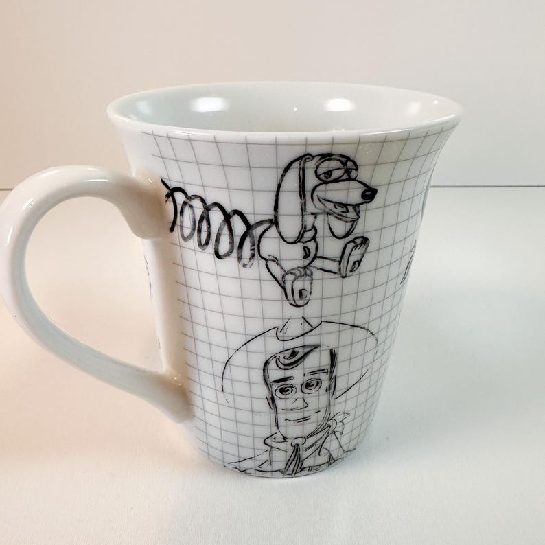 May include: White ceramic mug with black line drawings of Toy Story characters. The mug has a grid pattern and a curved handle. The illustrations include Slinky Dog and Woody. The mug is suitable for hot beverages.