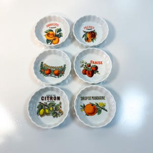Può includere: Sei piccoli piatti in ceramica bianchi con bordi smerlati e illustrazioni di frutta. Ogni piatto presenta un frutto diverso: albicocche, pesche, arance, fragole, limoni e un mandarino. Il testo in francese accompagna ogni frutto.