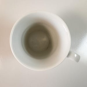 May include: Overhead shot of a white ceramic mug with a handle. The inside of the mug has a slightly darker, off-white hue. The mug is set against a white background.