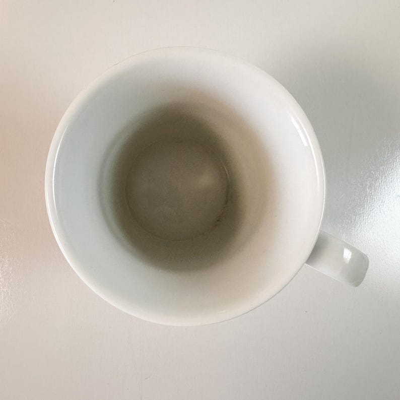 May include: Overhead shot of a white ceramic mug with a handle. The inside of the mug has a slightly darker, off-white hue. The mug is set against a white background.