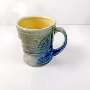 Puede incluir: Taza de cerámica hecha a mano con una forma orgánica. La taza presenta tonos azules, verdes y beige, con un interior amarillo. Un diseño de hojas azules adorna el lateral, y el asa es azul oscuro. Una pieza funcional y artística.