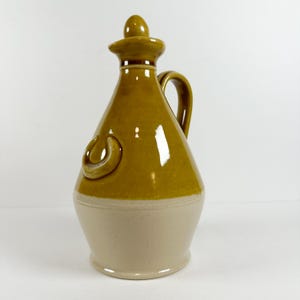 May include: A ceramic pitcher with a brown glaze and a white base. The pitcher has a handle and a spout. It has a unique design with a curved handle and a small spout.