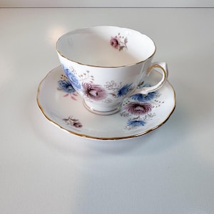 May include: White teacup and saucer set with gold trim. The cup and saucer are decorated with blue, pink, and purple floral designs. The cup has a curved handle and a scalloped rim. A classic tea set.