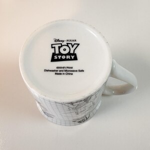 May include: White ceramic mug featuring the "Toy Story" logo and character sketches. The mug is dishwasher and microwave safe, made in China. The handle is white and the base of the mug is visible.