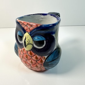 Puede incluir: Una taza de cerámica con forma de búho, con un cuerpo azul oscuro, ojos grandes y expresivos y un pico amarillo. La taza tiene un asa y está decorada con detalles rosas, azules y marrones. El pecho del búho tiene un patrón de escamas.