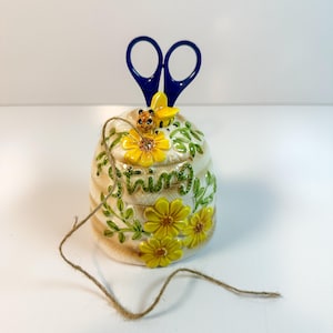 Puede incluir: Un recipiente de cerámica con forma de colmena con la palabra "Spring" en escritura verde, adornado con flores amarillas y una abeja. Tijeras azules se insertan en la parte superior. Un trozo de cuerda está sobre el frente.