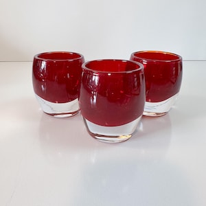 May include: Three red glass tumblers with a clear base are arranged on a white surface. The tumblers have a rounded shape and a glossy finish. The red color is rich and vibrant, contrasting with the clear base.