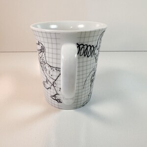 May include: White ceramic mug featuring a grid pattern and black line drawings of cartoon dinosaurs. The mug has a white handle and a slightly flared shape.