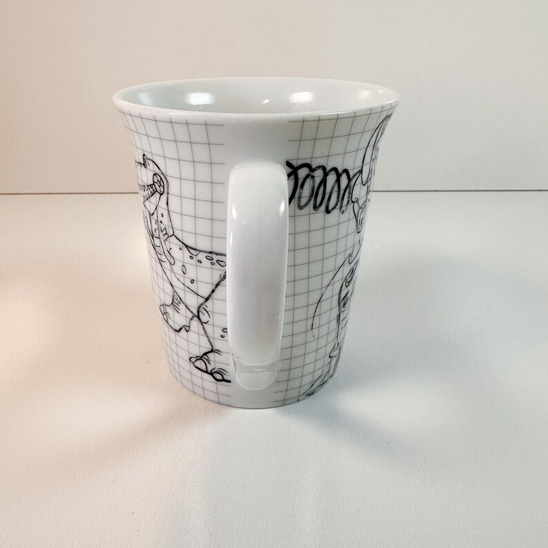May include: White ceramic mug featuring a grid pattern and black line drawings of cartoon dinosaurs. The mug has a white handle and a slightly flared shape.