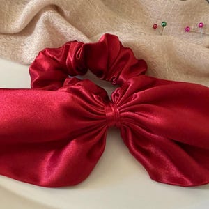 Peut inclure: Un chouchou en satin rouge vif avec un grand nœud. L'accessoire pour cheveux est fait d'un tissu lisse et brillant. Il est présenté sur une surface blanche avec un tissu beige en arrière-plan. Un accessoire de cheveux élégant.