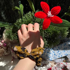 Peut inclure: Un chouchou en tissu avec un motif abstrait jaune moutarde et noir, agrémenté d'un nœud décoratif. L'accessoire est porté au poignet, mettant en valeur la texture froncée du tissu et la forme distincte du nœud sur un fond végétal.