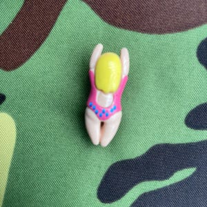 May include: A small plastic figurine of a person in a pink swimsuit with blue dots. The figurine has yellow hair and is in a diving pose.