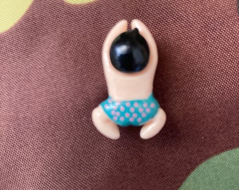 May include: A small ceramic figurine of a person in a blue and pink polka dot swimsuit with arms raised above their head. The figurine has a black head and a light pink body.