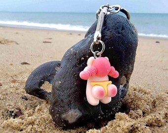 May include: A pink and yellow cartoon character keychain with a pink bow on its head. The keychain is hanging from a silver chain and is resting on a dark gray rock.