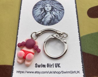 May include: A pink and red polymer clay keychain charm of a girl in a swimsuit. The charm is attached to a silver keyring with a spring clasp. The keychain is packaged on a white card with the text "Swim Girl UK" and a website address.