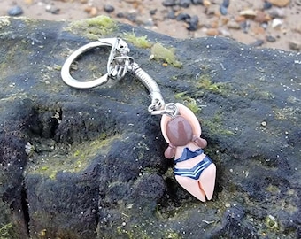 May include: A silver keychain with a miniature figurine of a person in a swimsuit. The figurine is brown with a blue and white striped swimsuit. The keychain is attached to a coiled metal piece and a silver ring.