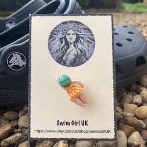 May include: A small, hand-painted ceramic pin of a swimmer wearing a yellow and red swimsuit and a blue and green swim cap. The pin is on a white card with the text "Swim Girl UK" and a website address.