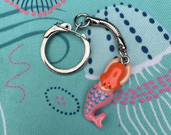 May include: A mermaid keychain with a silver keyring and chain. The mermaid has a pink body, blue scales, and orange hair. The keychain is on a teal background with a pink and blue pattern.