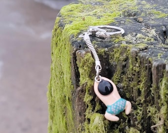 May include: A silver keychain with a baby charm. The charm features a cream-colored baby with a black head and blue diaper. The keychain is hanging on a moss-covered wooden post near the ocean.