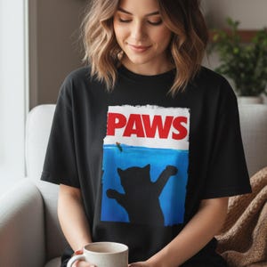 May include: Black t-shirt with a graphic design featuring the word "PAWS" in red letters above a blue ocean scene with a black cat silhouette reaching for a fish. The shirt is made of a soft material.