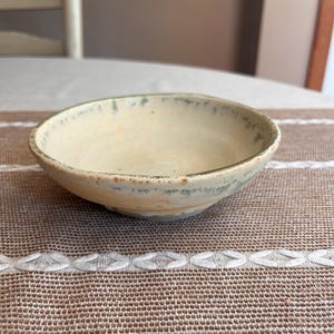 May include: A shallow, circular ceramic bowl with a cream-colored interior and a textured, mottled rim featuring shades of sage green and earthy brown. The exterior surface shows subtle variations in tone, with a smooth, slightly uneven finish.