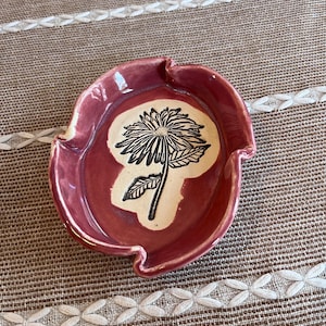 May include: A small, oval-shaped ceramic dish with a glossy, deep rose-colored glaze. The center features a cream-colored inlay decorated with a detailed, black-inked illustration of a single daisy-like flower with leaves and a stem.