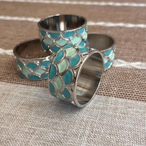 May include: A set of four circular napkin rings featuring a metallic silver finish and an exterior pattern of overlapping scales in alternating shades of teal and light mint green. The interior of each ring is smooth, polished silver-tone metal.