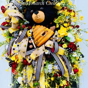 May include: A decorative wreath featuring a black bear head, a beehive, and various ribbons. The wreath includes yellow, red, and white flowers, along with butterflies and berries. The text "Black Monarch Creations" is visible.